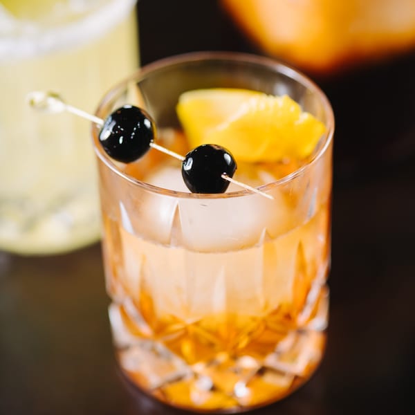 A Thailicious cocktail with ice, an orange slice, and two cherries on a skewer.