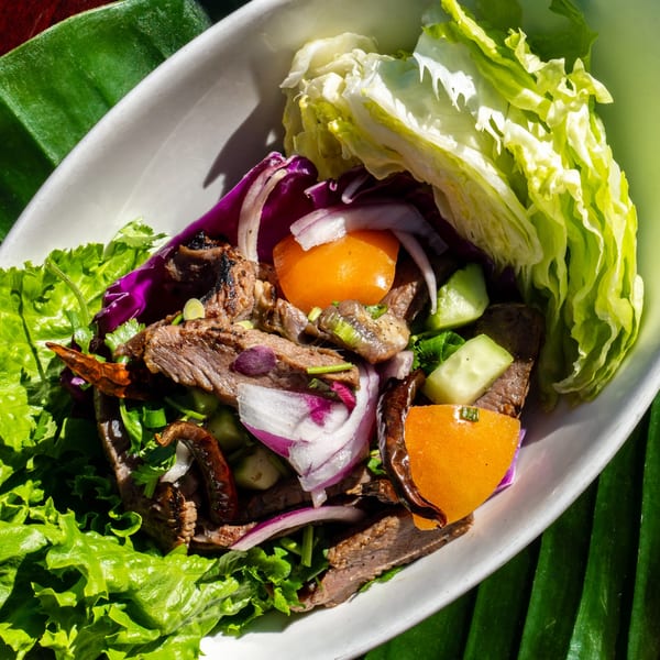 Thailicious Thai beef salad with lettuce, red onion, tomatoes, and cucumbers in a white bowl.