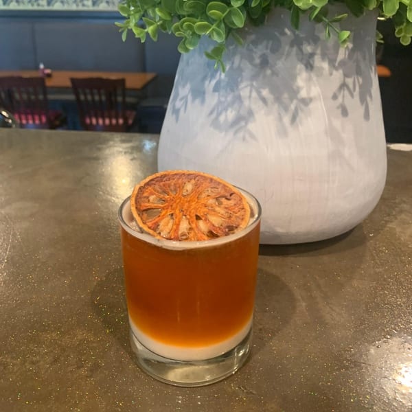 A Thailicious cocktail with a dried orange slice, next to a white vase with greenery.